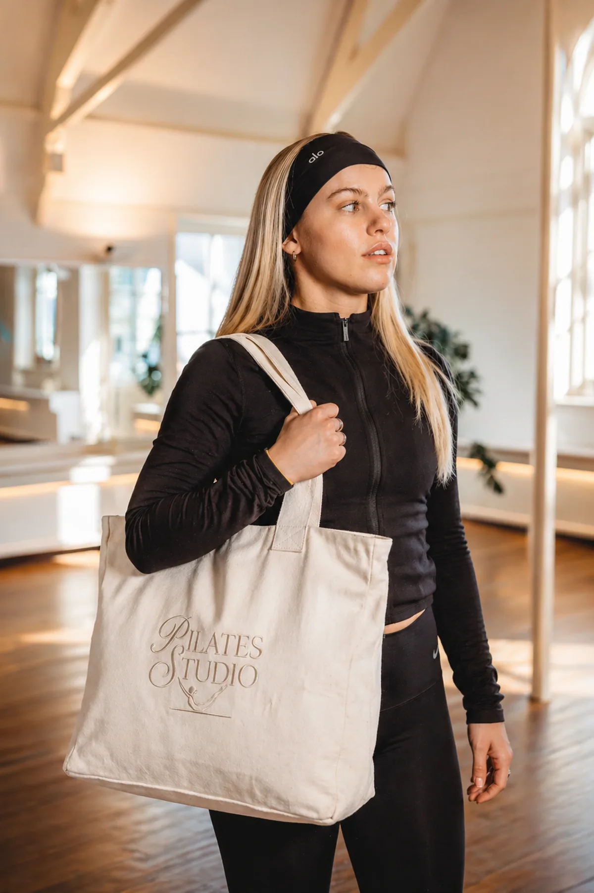 Pilates studio member carrying custom branded canvas tote bag with embroidered Pilates Studio logo in bright reformer studio